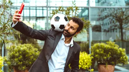 Small business owner balancing soccer ball on shoulder while taking selfie outdoors, representing creative marketing during a global sports event