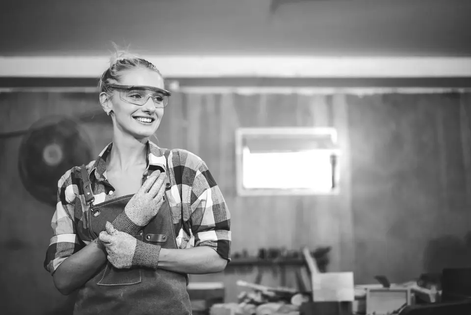 A handyman smiles and wears safety glasses and overalls in a work shop